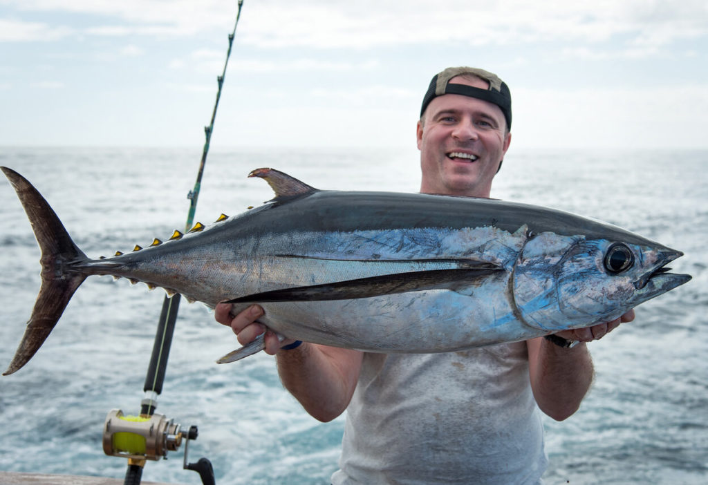 Ein fröhlicher Angler, der auf einem Boot einen Blauflossen-Thunfisch in die Kamera hält.